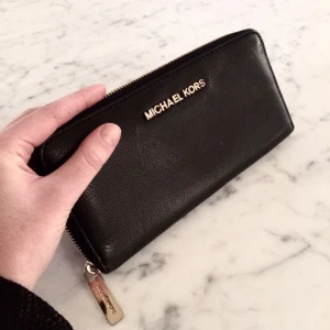  - Plånbok från Michael Kors. Modell Jet set Travelza. Svart läder och guldfärgade detaljer. Nypris 1400 kr. Litet slitage på bokstäverna, se bild 2.