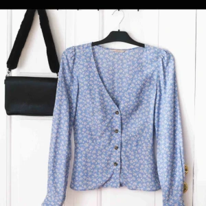  - En fin, figursydd blus i en härlig blå färg med blommönster och knappar framtill. Det finns en söm i midjan som ger en minimal peplum- effekt och framhäver midjan på fint sätt. 