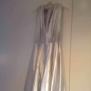  - Lång balklänning i silvertyg, halterneck och långa band som hänger ner över ryggen. 