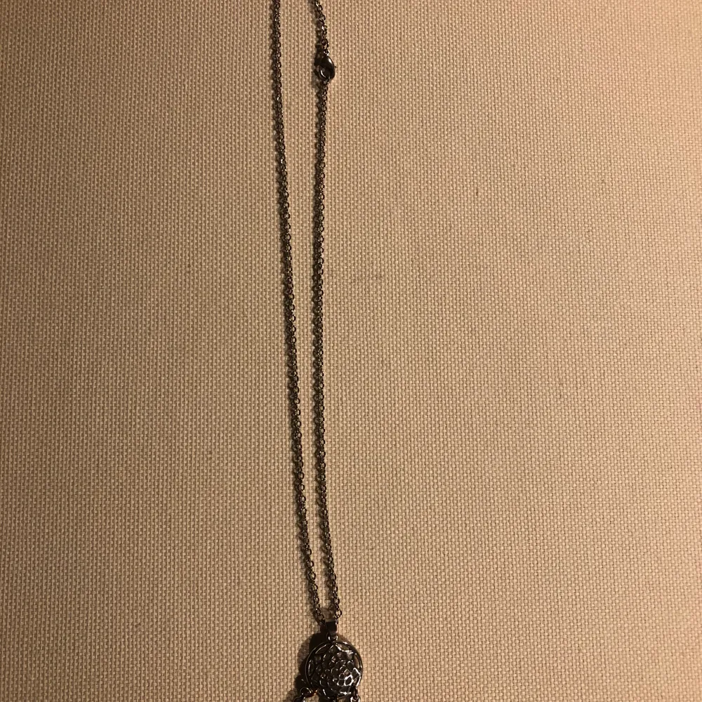 Ett silverfärgat halsband med en liten drömfångare som berlock. Asusteet.