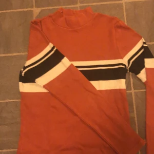 Polotröja  - Tight långärmad tröja med polo krage. Tröjan går i en terracotta färg med ränder i svart och vitt. 