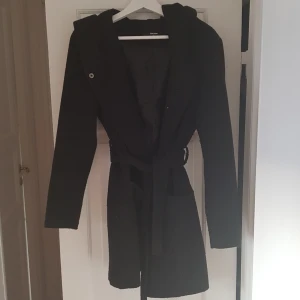  - Jacka från BikBok, svart med skärp i midjan, två stora fickor framtill, stor luva, knäppa med dolda knappar, halvlång, för höst/vinter. Knäppt använd. Kostar 800 kr ny.