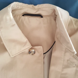  -  Trench coat, herr. Studio Mario Bigotti. Khaki med svarta detaljer och ljust foder. Två yttre fickor och två innerfickor med dragkedja. Storlek S/M, Längd 100 cm, axelbredd 50 cm, ärm 60 cm. Ny, oanvänd. Nypris ca 2.000 kr. Säljes 500 kr.