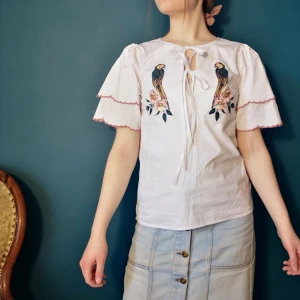 Papegojblus - Vit blus med broderade papegojor, snörning och volangärmar 🦜🌺 Aldrig använd. Frakt tillkommer! 