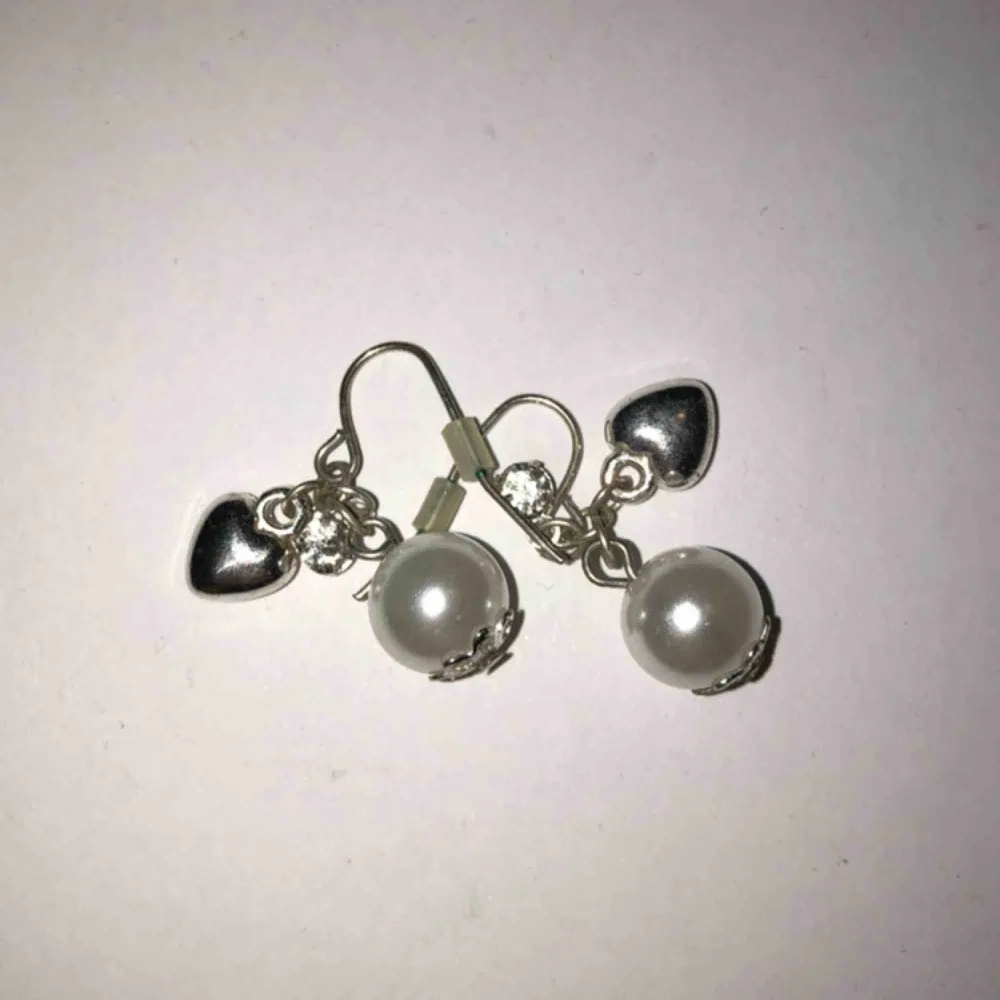 Ett par hängande örhängen med en vit pärla, ett hjärta och en kristall. Helt oanvända. Köparen betalar frakten. Accessoarer.
