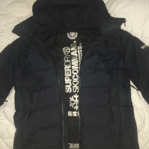 Varm Superdry vinterjacka i storlek M - Praktisk vinterjacka med extra-fickor på båda ärmarna. Håller värmen utan problam samtidigt som man kan vädra under armarna ifall det skulle bli för varmt, skulle funka bra till en skidresa men funkar annars suveränt som en vanlig vinterjacka. Strl: M