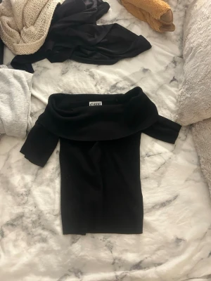 Svart offshoulder topp - Snygg svart offshoulder topp med bred krage och korta ärmar. Toppen har en figurnära passform och är perfekt för dig som vill ha en stilren och trendig look. Materialet känns mjukt och stretchigt, vilket gör den bekväm att bära.