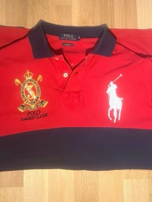 Röd piké från Polo Ralph Lauren - Säljer en röd pikétröja från Polo Ralph Lauren med marinblå krage och detaljer. Stor logga med polospelare på bröstet och broderad gul detalj med texten 'Polo Summer Classic'. Klassisk custom fit-modell.