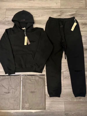 Svart Essentials hoodie med huva - Säljer en svart hoodie från Essentials med diskret logga på bröstet. Hoodien har en klassisk passform, stor känguruficka framtill och ribbade muddar vid ärmar och midja. Tillverkad i mjukt och bekvämt material, perfekt för en avslappnad streetwear-look.