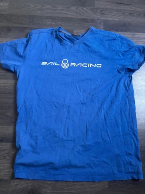 Blå Sail Racing t-shirt - Snygg blå t-shirt från Sail Racing med vit logga tryckt över bröstet. Klassisk rund halsringning och korta ärmar. Tillverkad i mjuk bomull som känns skön mot huden. Perfekt för en avslappnad och sportig stil.