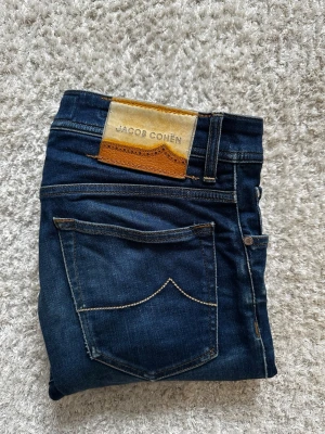 Mörkblå jeans från Jacob Cohën - Snygga mörkblå jeans från Jacob Cohën med klassisk femficksmodell och kontrastsömmar. Jeansen har en skinnpatch med logga bak i midjan och smal passform. Perfekta för dig som gillar stilrena och tidlösa jeans i mjukt denimtyg. Se sista bild för ett 3-4mm hack. 