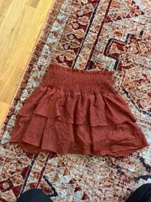 Vinröd volangkjol med guldiga glitter-detaljer - Kort brun kjol från Guts & Gusto med volanger och små guldprickar. Kjolen har bred smock i midjan för en bekväm passform och är perfekt för dig som gillar en lekfull och trendig stil.