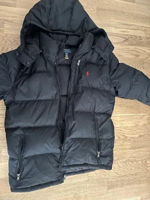 Svart dunjacka från Polo Ralph Lauren - Säljer en svart dunjacka från Polo Ralph Lauren med röd logga på bröstet. Jackan har huva, dragkedja framtill och två sidofickor med dragkedja. Perfekt för kalla vinterdagar och riktigt skön att bära med inga skador, köpt förra vintern.