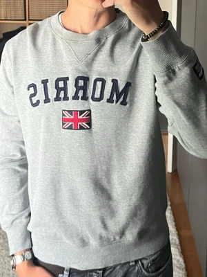 Morris sweatshirt med logga i grå - stl M - Morris sweatshirt med logga i gråmelerad, storlek M. 100% Bomull. Broderad logga och flagga fram, märke på ärmen. Ruggad insida, tjock och varm. Personen på bilden är 189cm, väger 78kg.  Använd men i mycket fint skick, inga hål eller fläckar.  Rök- och djurfritt hem. Skriv gärna om du vill ha fler bilder eller undrar något!