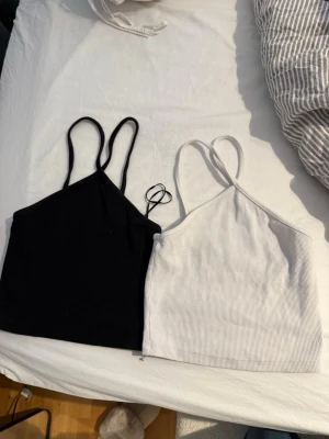 Svart och vit ribbad linnetopp - Två stilrena linnetoppar i svart och vitt med smala axelband och ribbad struktur. Perfekta att matcha till jeans eller kjol för en enkel och trendig look. Båda topparna är ärmlösa och har en croppad passform.