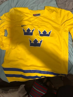 Gul Tre Kronor hockeytröja Nike - Säljer en klassisk gul hockeytröja från Nike med Tre Kronor-tryck på bröstet och blå detaljer. Tröjan har lång ärm och är perfekt för dig som vill stötta Sverige på match eller träning. Luftigt material och sportig passform.
