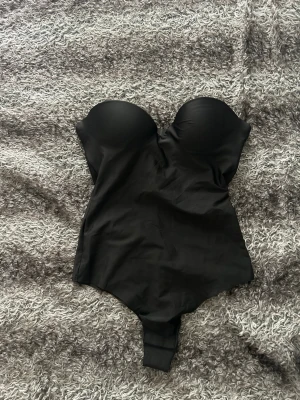 Svart body med avtagbara axelband - Snygg svart body med vadderade kupor och avtagbara axelband. Kan bäras både axelbandslös och med tunna band. Mjuk och stretchig känsla, perfekt för en stilren look under kläderna.