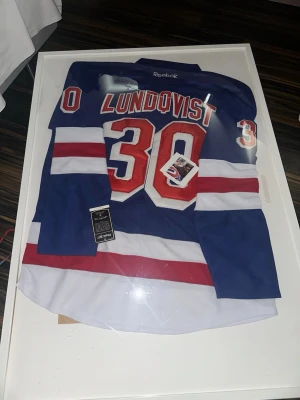 Blå Reebok NHL-tröja Lundqvist med hans autograf  - Säljer en blå NHL-hockeytröja från Reebok med namn Lundqvist och nummer 30 i rött och vitt med hans autograf och bevis. Tröjan har vita och röda detaljer på ärmar och nederkant. Perfekt för hockeyfans och samlare!