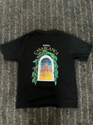 Svart Casablanca T-shirt - Snygg svart t-shirt av märket Casablanca med tryck av en färgglad port och texten 'Avenida Casablanca' både fram och bak. T-shirten har korta ärmar och normal passform. Perfekt för dig som gillar grafiska prints och streetwear-vibe.