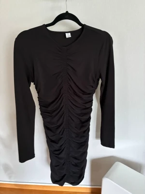 Svart långärmad klänning  - Säljer en svart långärmad klänning med rynkade detaljer framtill. Klänningen har en kroppsnära passform och är tillverkad i ett mjukt, stretchigt material. Perfekt för dig som gillar stilrena och trendiga plagg.