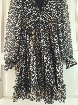 Vero moda small Leopardmönstrad volangklänning - Kort klänning med leopardmönster i svart, beige och bruna toner. Klänningen har v-ringning, långa ärmar och flera volanglager som ger en luftig känsla. Tillverkad i ett lätt, transparent material med resår vid ärmsluten. 
