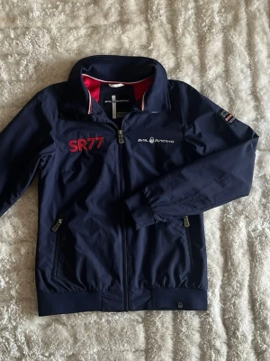 Sail Racing SR77 seglarjacka marinblå - stl M - Snygg marinblå jacka från Sail Racing med SR77 detaljer och klassisk seglarstil. Jackan har dragkedja framtill, två sidofickor med dragkedja och ribbad mudd nedtill och vid ärmsluten. Tydliga SR77 och Sail Racing loggor samt detaljer på ryggen.  Storlek M, men upplevs något mindre i storleken passar ungefär som en mindre M. Använd men i mycket fint skick, inga hål eller fläckar.  Rök- och djurfritt hem. Skriv gärna om du vill ha fler bilder eller undrar något!