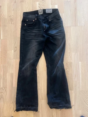 Svarta bootcut jeans med rå kant - Säljer ett par svarta bootcut jeans med rå, fransig kant nedtill. Jeansen har fem fickor, silverfärgade nitar och en lätt tvättad look. Passar perfekt till en avslappnad och trendig stil. Materialet är denim med stretch.