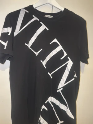 Svart t-shirt med stor vit logga - Snygg svart t-shirt med stor vit VLTN-logga tryckt diagonalt över hela framsidan. Klassisk rund hals och korta ärmar. Perfekt för dig som gillar statement-plagg och vill sticka ut lite extra.