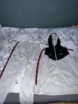 Nike grått tracksuit med huva - Nike tracksuit i ljusgrått med svarta och röda detaljer. Setet består av zip-hoodie med svart ovandel, vit nederdel och huva samt matchande byxor. Båda delarna har Nike-logga och röda band med vita swoosh längs ärmar och ben. Perfekt för träning eller chill. 2 Små märken.