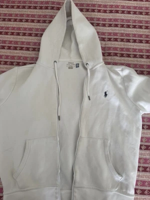 Vit hoodie från Polo Ralph Lauren - Snygg vit hoodie från Polo Ralph Lauren med dragkedja, två fickor framtill och klassisk huva med dragsko. Ikoniska lilla loggan broderad på bröstet. Tillverkad i mjukt bomullsmaterial som känns skönt mot huden.