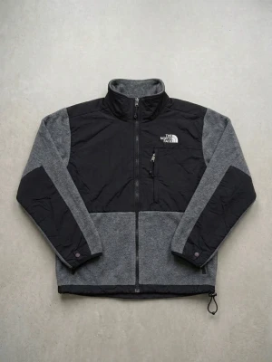 Grå och svart fleecejacka The North Face - Snygg grå och svart fleecejacka från The North Face med dragkedja framtill, hög krage och bröstficka med logga. Jackan har justerbar nederkant och förstärkta partier på axlar och ärmar för extra slitstyrka.