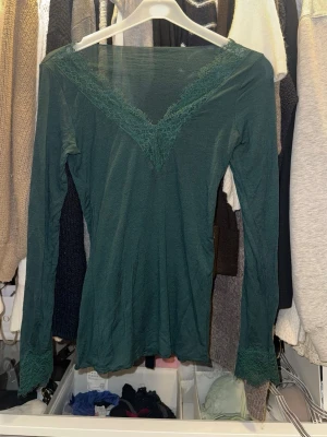 Mörkgrön långärmad topp med spets - Mörkgrön långärmad topp med v-ringning och dekorativ spets längs halsringningen och vid ärmsluten. Tunn och mjuk känsla, perfekt för lager på lager eller som den är. Snygg och enkel att matcha med olika stilar.