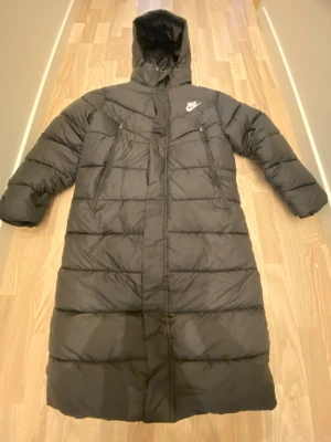 Svart lång dunjacka från Nike - Säljer en svart lång dunjacka från Nike med huva, dragkedja och knappar hela vägen ner. Jackan har två sidofickor med dragkedja och en diskret Nike-logga på bröstet. Perfekt för kalla vinterdagar och riktigt skön att bära.