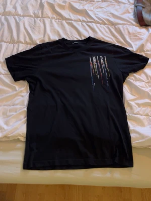 Svart AMIRI t-shirt med färgstänk - Säljer en svart t-shirt från AMIRI med tryck av loggan och färgstänk i flera färger både fram och bak. T-shirten har korta ärmar och en klassisk passform. Perfekt för dig som gillar streetwear och vill sticka ut med en unik design.
