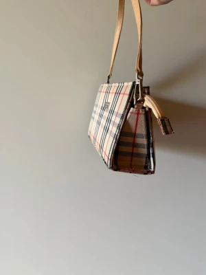 Rutig handväska med beige axelrem - Trendig handväska i klassiskt rutigt mönster med beige, svart och röda detaljer. Väskan har en rak, boxig form och beige axelrem i läderimitation. Har ärvt den därav priset.