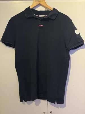 Svart Moncler pikétröja - Snygg svart pikétröja från Moncler med klassisk krage och korta ärmar. Tröjan har en liten knappslå framtill och Monclers ikoniska logga på vänster ärm. Perfekt till jeans eller shorts för en clean och stilren look.