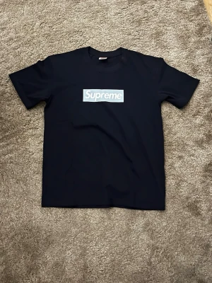Supreme x Tiffany & Co svart t-shirt - Supreme x Tiffany & Co svart t-shirt med klassisk box logo framtill i ljusblått och vitt. På ryggen finns Tiffany & Co-tryck i vitt. T-shirten har rund hals och korta ärmar, tillverkad i mjuk bomull för en clean och stilren look.