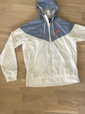 Nike vindjacka med huva, Helt ny!  - Nike vindjacka i vitt och ljusblått med huva och dragkedja. Jackan har meshfoder, två sidofickor och justerbara snören i huvan. Nike-logga i orange på bröstet ger en sportig vibe. Perfekt för blåsiga dagar och streetwear-stil. Jackan är helt ny, endast provad!!!