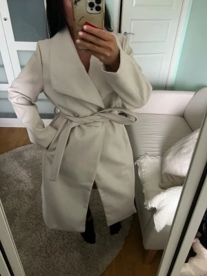 Beige lång kappa med skärp - Stilren kappa i perfekta begiea färgen. Kappan är rak i passformen och lång i benen. Perfekt för höst men funkar även på vintern eftersom den är ganska varm. Den är bara använd typ 3 gånger och väldigt varsamt.
