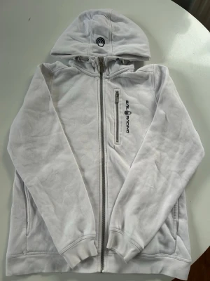 Vit hoodie från Sail Racing med dragkedja - Vit hoodie från Sail Racing med hel dragkedja framtill, huva och en bröstficka med dragkedja. Logga på bröstet och på huvan. Mjuk bomullskänsla och ribbade muddar vid ärmslut och nederkant. Perfekt för en avslappnad och sportig stil.