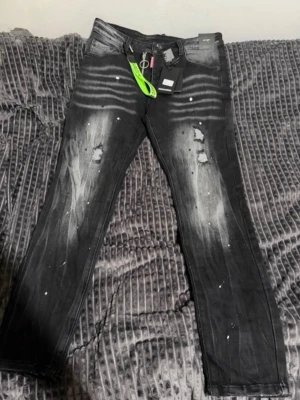 Svarta slitna jeans med detaljer - Säljer ett par svarta jeans med slitningar och vita färgstänk för en edgy look. Jeansen har raka ben, fem fickor och dragkedja med ringdetalj. Perfekta för dig som gillar streetwear och vill sticka ut lite extra.