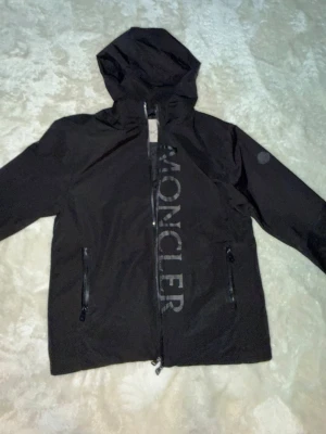 Svart vindjacka från Moncler - Snygg svart vindjacka från Moncler med huva och dragkedja framtill. Jackan har två fickor med dragkedja och ett stort Moncler-tryck längs dragkedjan. Tillverkad i ett lätt och vattenavvisande material, perfekt för blåsiga dagar.