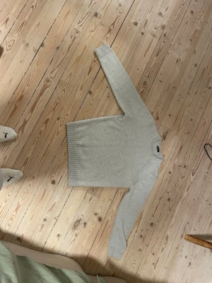 Ljusgrå basic stickad tröja - En enkel och stilren ljusgrå stickad tröja med rund hals och långa ärmar. Tröjan har ribbade muddar vid ärmslut och nederkant, vilket ger en klassisk look. Perfekt att matcha med jeans eller kjol för en clean outfit.
