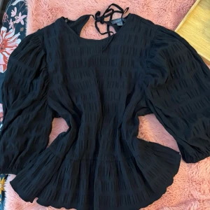 Svart blus med puffärmar och knyt - Trendig svart blus med puffiga långärmar och knyt i nacken. Blusen har en lätt strukturerad yta och en lös, flowy passform som ger en cool siluett. Perfekt att styla med jeans eller kjol för en snygg look. Den e storlek L men jag är storlek M och funkar jättebra 