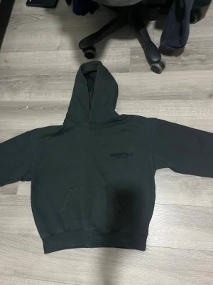 Svart Essentials Fear of God hoodie - Säljer en svart hoodie från Essentials Fear of God med stor huva och magficka. Tryck med 'ESSENTIALS' på bröstet och ryggen. Hoodien har en loose passform och är tillverkad i mjuk bomull, perfekt för en avslappnad streetwear-look.