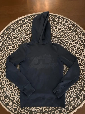 Mörkblå hoodie med tryck - Snygg mörkblå hoodie med stor huva och ett diskret tryck på bröstet. Hoodien har långa ärmar, ribbade muddar vid ärmslut och nederkant samt en klassisk passform. Perfekt för en avslappnad och cool stil.