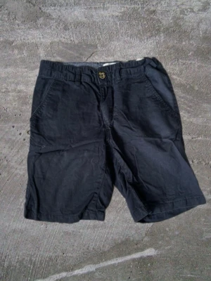 Mörkblå Shorts - Snygga mörkblå shorts i bomull med normal passform. Shortsen har två sidofickor fram, bälteshällor och knappstängning. På baksidan finns två fickor, varav en med en liten läderdetalj. Perfekta för varma dagar.