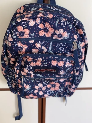 Blommig ryggsäck från Jansport - Mörkblå ryggsäck från Jansport med rosa och vita blommor. Flera fack med dragkedja, justerbara axelband och sidoficka i mesh. Perfekt för skola eller utflykter, rymmer mycket och har en snygg, ungdomlig design.