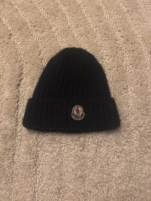 Moncler Mössa  - Snygg svart ribbstickad mössa från Moncler med uppvikt kant och klassisk Moncler-logga framtill. Perfekt för kalla dagar och enkel att matcha med olika outfits. Materialet ser ut att vara mjukt och värmande, troligen ull eller ullmix.