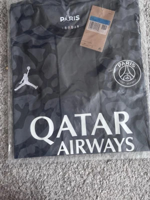 PSG x Jordan svart matchtröja - Snygg svart PSG matchtröja i samarbete med Jordan. Tröjan har ett camomönster, vita logotyper och texten 'Qatar Airways' på bröstet. PSG-emblemet och Jumpman-loggan syns tydligt framtill. Perfekt för fotbollsfans som vill sticka ut.